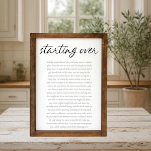 May include: Wooden framed sign with the words "starting over" in a handwritten font. The sign has a white background with black text, featuring song lyrics. The frame is a dark brown color.