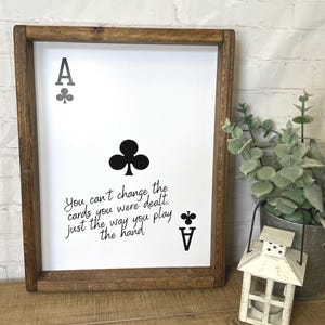 Puede incluir: Un cartel enmarcado con fondo blanco y texto negro. El cartel presenta un as de tréboles negro con la cita "No puedes cambiar las cartas que te han repartido. Solo la forma en que juegas la mano."