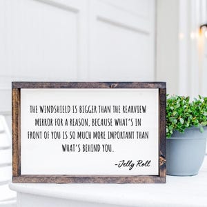 May include: A wooden framed sign with the quote "THE WINDSHIELD IS BIGGER THAN THE REARVIEW MIRROR FOR A REASON, BECAUSE WHAT'S IN FRONT OF YOU IS SO MUCH MORE IMPORTANT THAN WHAT'S BEHIND YOU." The sign is signed "-Jelly Roll."
