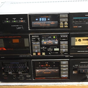 May include: A stack of three vintage black stereo components. The top unit is a tuner with a digital display showing 99.5 FM. The middle unit is a cassette deck, and the bottom unit is an amplifier.