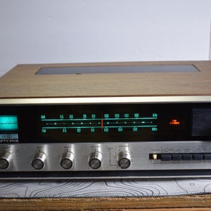 May include: A Model Fifty-Five stereo receiver with a wood grain top and a black front panel. It features a green illuminated AM/FM radio dial, a signal strength meter, five silver control knobs, a row of black push buttons, and a headphone jack.