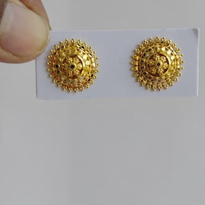 Può includere: Un paio di orecchini a lobo dorati e rotondi caratterizzati da un design testurizzato ispirato al sole, con una cupola centrale rialzata e intricati dettagli a perline lungo i bordi, montati su un cartoncino bianco rettangolare.