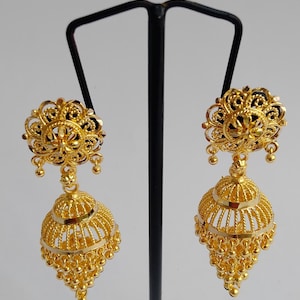 May include: A pair of gold-coloured, ornate earrings. Each earring features a floral design at the top, a bell-shaped body with intricate detailing, and small dangling beads. The earrings are displayed on a black stand.