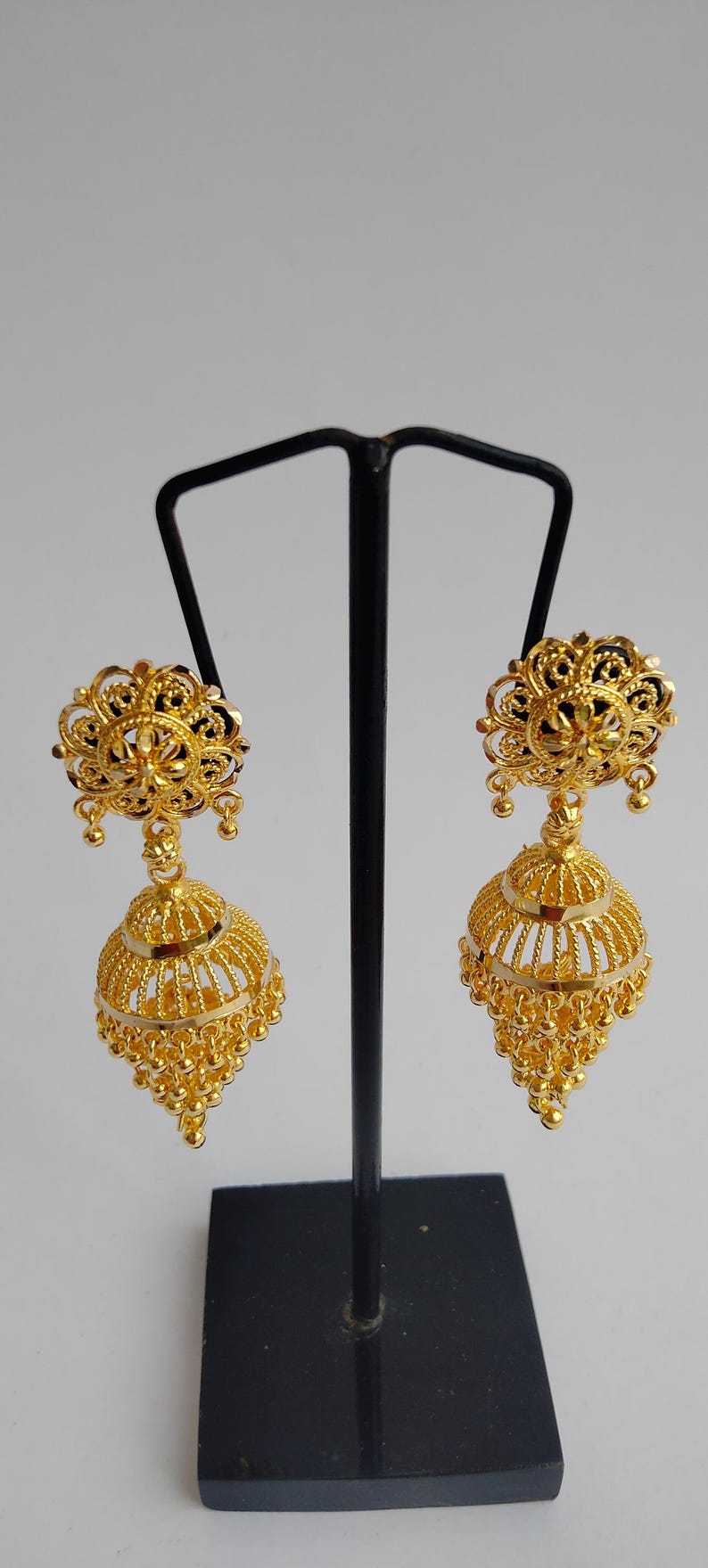May include: A pair of gold-coloured, ornate earrings. Each earring features a floral design at the top, a bell-shaped body with intricate detailing, and small dangling beads. The earrings are displayed on a black stand.