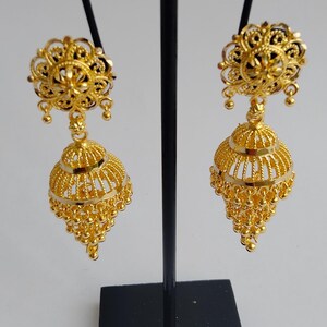 May include: A pair of gold-coloured, ornate earrings. Each earring features a floral design at the top, connected to a bell-shaped pendant with intricate filigree and small dangling beads. The earrings are displayed on a black stand.