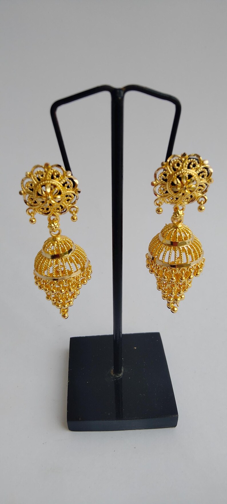 May include: A pair of gold-coloured, ornate earrings. Each earring features a floral design at the top, connected to a bell-shaped pendant with intricate filigree and small dangling beads. The earrings are displayed on a black stand.