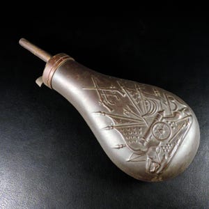 May include: Antique metal powder flask with a detailed embossed design of a sailing ship, cannons, and flags. The flask has a dark, aged appearance with a spout and a small metal loop. The background is black.
