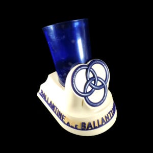 May include: A vintage Ballantine Ale bar caddy with a blue glass and a cream-colored base. The base features the Ballantine Ale logo in blue and a three-ring design. The caddy is set against a black background.