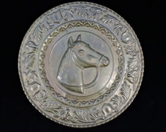 May include: A round, brass wall plaque with an ornate design featuring a horse's head in the center. The horse is in relief and has a bridle.