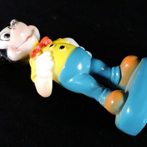 May include: A vintage figurine of a cartoon character, lying on its back. The figure has a white face, a red bow tie, a yellow shirt, blue trousers, and orange shoes. The figure is lying on a black surface.