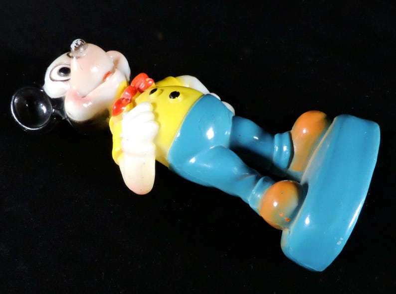 May include: A vintage figurine of a cartoon character, lying on its back. The figure has a white face, a red bow tie, a yellow shirt, blue trousers, and orange shoes. The figure is lying on a black surface.