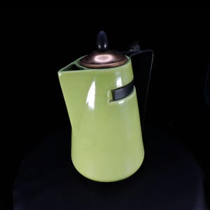 May include: A lime green ceramic teapot with a tapered body and a wide spout. It features a metallic copper-toned lid topped with a black finial. A thin black metal handle is attached to the side, and a black horizontal band accents the upper body.