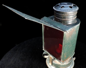 May include: A vintage green metal lantern with a red glass panel and a hinged lid. The lantern has a black metal top with a circular opening and a small handle.
