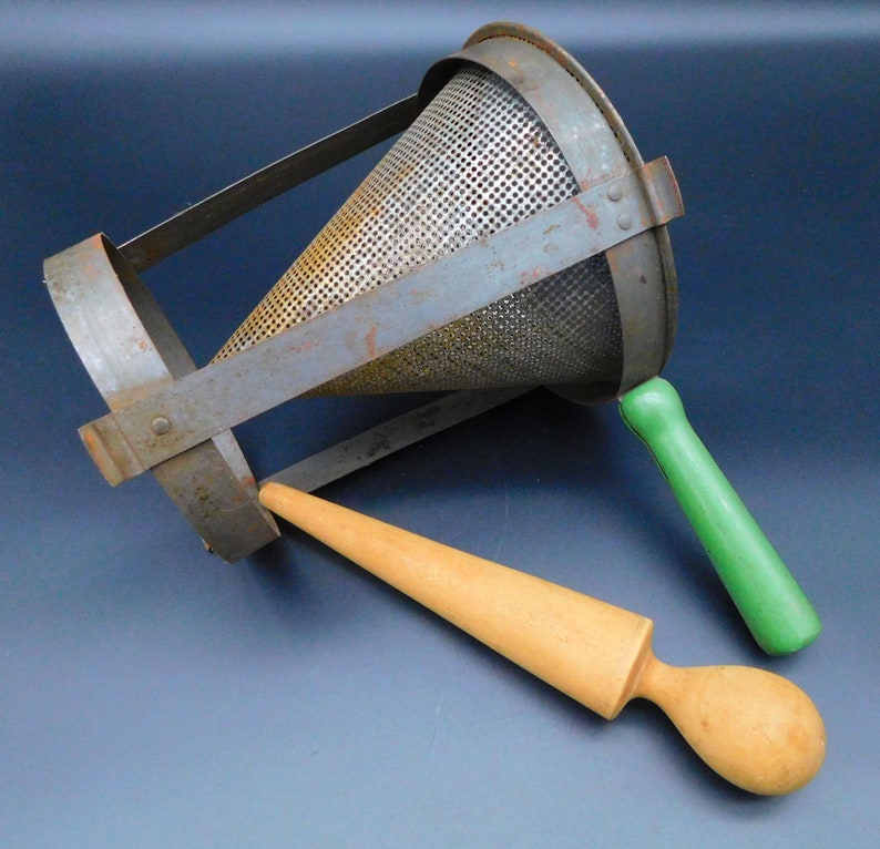 May include: A vintage metal potato ricer with a green wooden handle and a wooden pestle. The ricer is made of metal with a mesh design and has a handle that is attached to the side. The pestle is made of wood and has a rounded end.