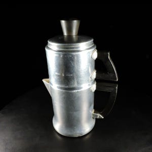 May include: A vintage aluminum coffee percolator with a black handle and lid. The coffee pot has a cylindrical body with a spout and a black handle. The lid is also black. The percolator is set against a black background.