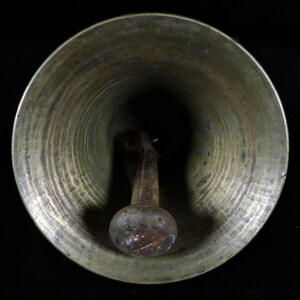 May include: A bronze-colored bell with a rounded bottom and a clapper. The bell's interior is dark, and the clapper has a bulbous end. The bell has a slightly aged appearance, suggesting it may be vintage or antique.