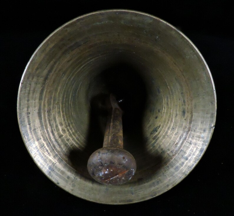 May include: A bronze-colored bell with a rounded bottom and a clapper. The bell's interior is dark, and the clapper has a bulbous end. The bell has a slightly aged appearance, suggesting it may be vintage or antique.
