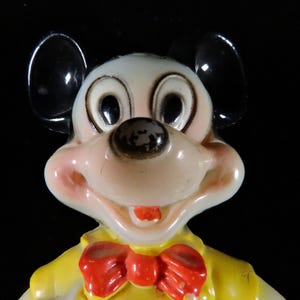 May include: A vintage Mickey Mouse figurine. The figure has a white face, black ears, and a red bow tie. The figure is wearing a yellow jacket. The figurine is a collectible item.