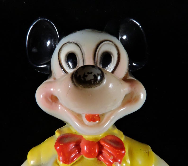 May include: A vintage Mickey Mouse figurine. The figure has a white face, black ears, and a red bow tie. The figure is wearing a yellow jacket. The figurine is a collectible item.