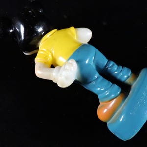 May include: A vintage Mickey Mouse figurine, with a black head, yellow shirt, blue trousers, and orange shoes. The figure is in a playful pose, with one foot extended. The toy is made of a glossy material.