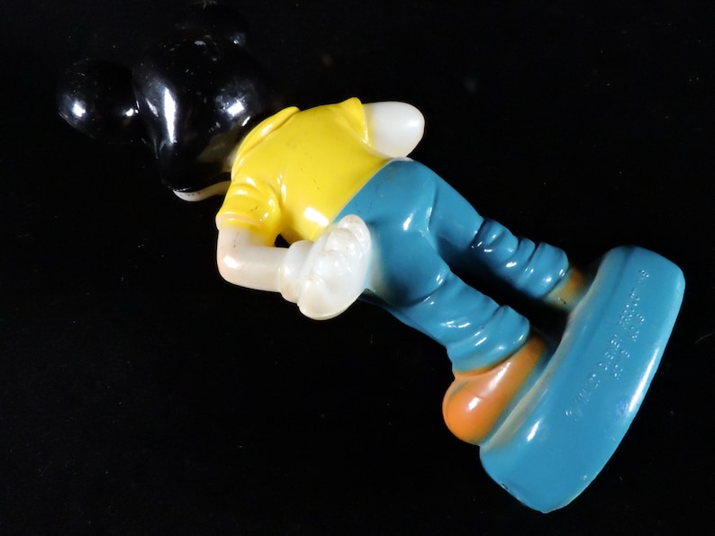 May include: A vintage Mickey Mouse figurine, with a black head, yellow shirt, blue trousers, and orange shoes. The figure is in a playful pose, with one foot extended. The toy is made of a glossy material.