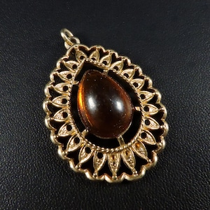 May include: A gold-toned pendant with a teardrop-shaped amber-colored stone. The pendant has an ornate, openwork design with a scalloped edge and a twisted rope border around the stone. The pendant is on a black surface.