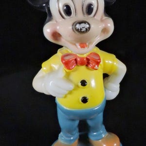 May include: A vintage Mickey Mouse figurine. The figure is wearing a yellow shirt with black buttons, blue trousers, and orange shoes. A red bow tie and white gloves complete the outfit. The figurine stands on a blue base.