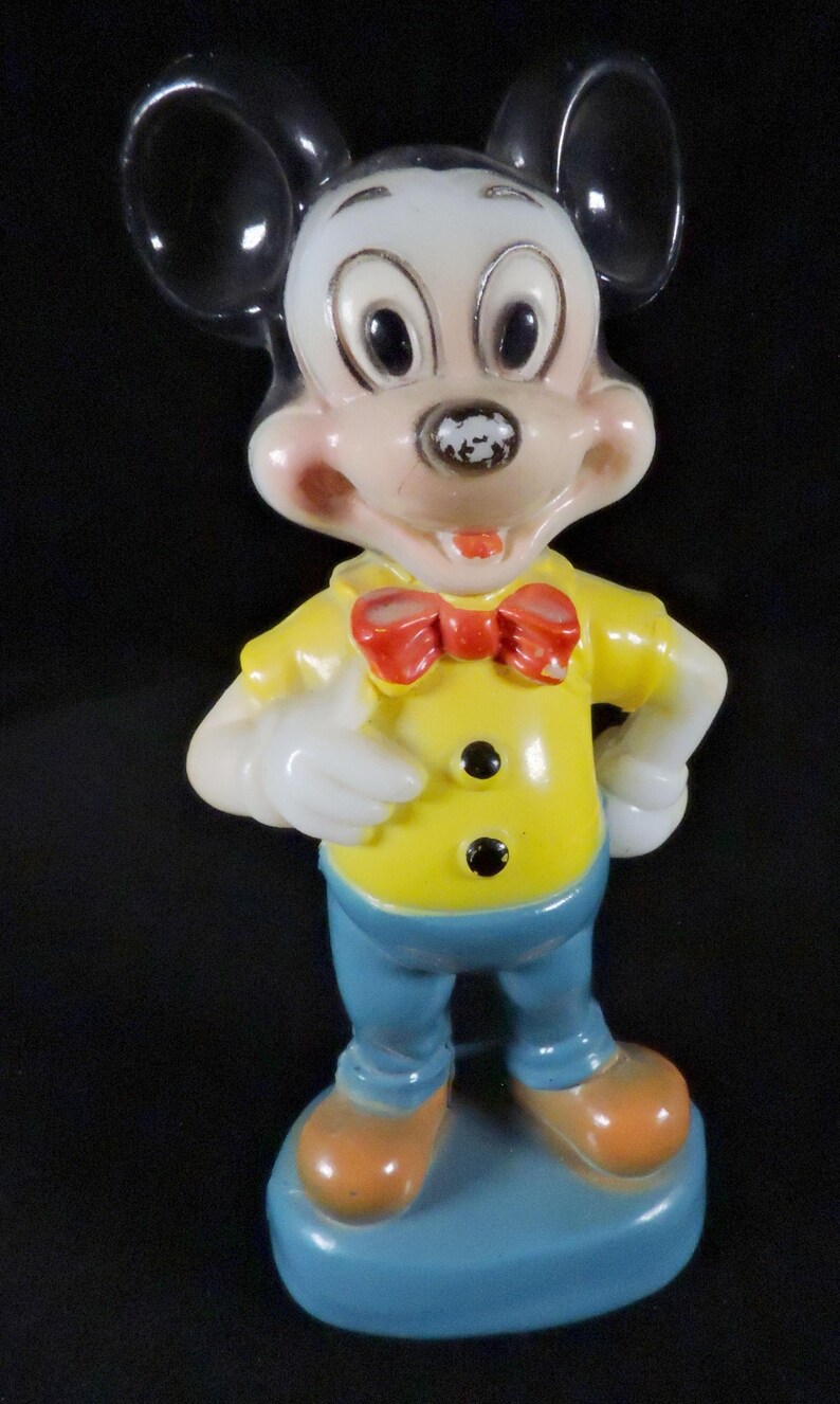May include: A vintage Mickey Mouse figurine. The figure is wearing a yellow shirt with black buttons, blue trousers, and orange shoes. A red bow tie and white gloves complete the outfit. The figurine stands on a blue base.