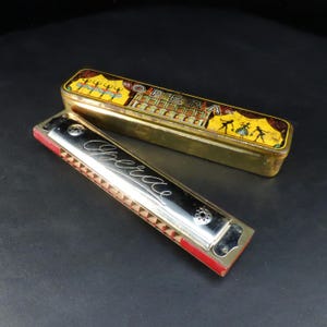 May include: A silver-tone harmonica with the word Opera engraved in cursive script, featuring red side panels and circular air holes. Beside it lies a rectangular gold-tone metal case decorated with a yellow illustration of dancers and the word Opera.