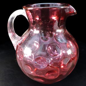 May include: A vintage pink glass jug with a clear handle and spout. The jug features a pattern of raised circles across its surface. The glass has a glossy finish and reflects light, creating highlights and shadows.