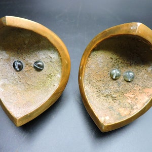 May include: Two brass candle holders with a heart shape and a textured surface. Each holder has two small, round, black and white marble-like accents.