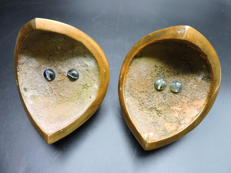 May include: Two brass candle holders with a heart shape and a textured surface. Each holder has two small, round, black and white marble-like accents.