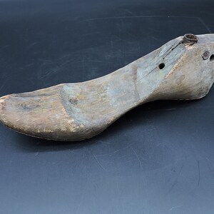 May include: A wooden shoe last, a tool used by shoemakers to shape and form shoes. The last is made of light brown wood and has a worn, aged appearance. It has several holes and a metal piece at the top.