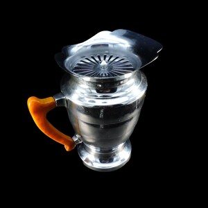 May include: A polished silver-tone metal pitcher featuring a wide, flared rim with an integrated circular strainer insert. The vessel has a rounded body and a distinct, curved handle in a solid, opaque amber-colored material attached to the side.