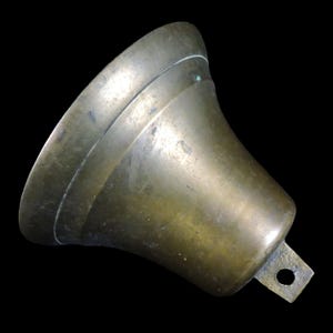 May include: A bronze-colored bell with a flared opening and a loop for hanging. The bell has a slightly weathered appearance, with a dark background. The bell's design includes a prominent horizontal ridge near the top.