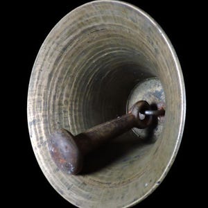 May include: A close-up view inside a bronze-colored bell. The bell has a flared shape with concentric circular ridges. A rusty, cylindrical clapper hangs inside, attached by a metal loop. The background is black.