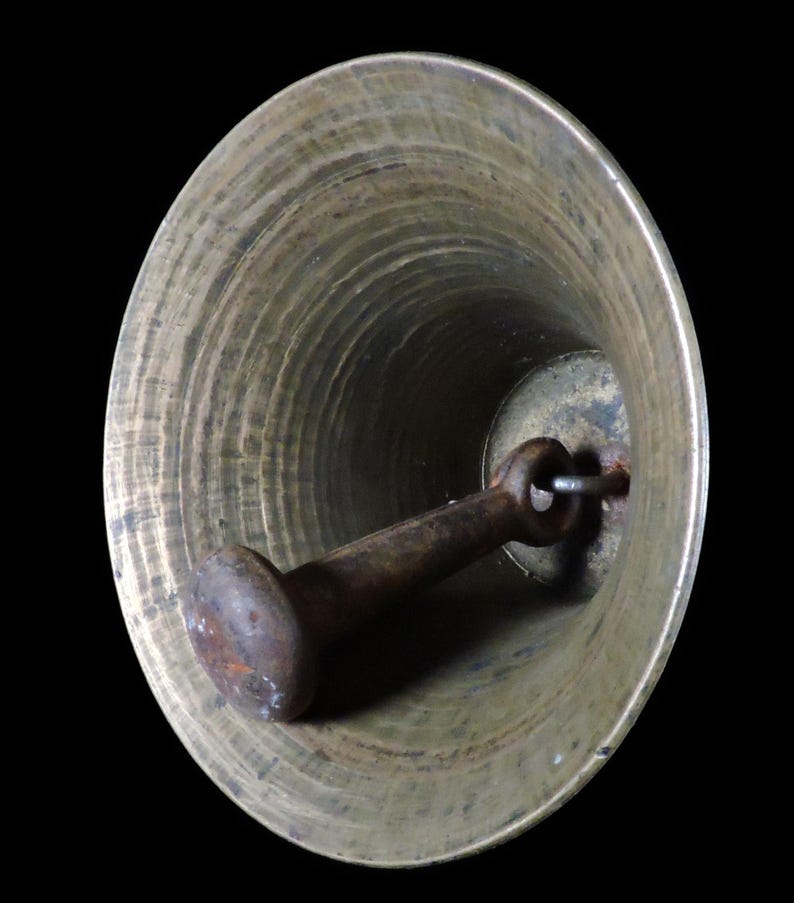 May include: A close-up view inside a bronze-colored bell. The bell has a flared shape with concentric circular ridges. A rusty, cylindrical clapper hangs inside, attached by a metal loop. The background is black.