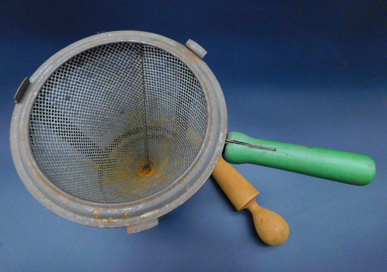 May include: A vintage metal strainer with a green and wooden handle. The strainer has a mesh bottom and a cone shape. The handle is attached to the side of the strainer.