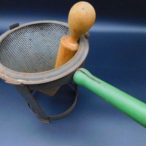 May include: A vintage metal food mill with a green wooden handle and a wooden pestle. The mill has a mesh strainer and a metal base.