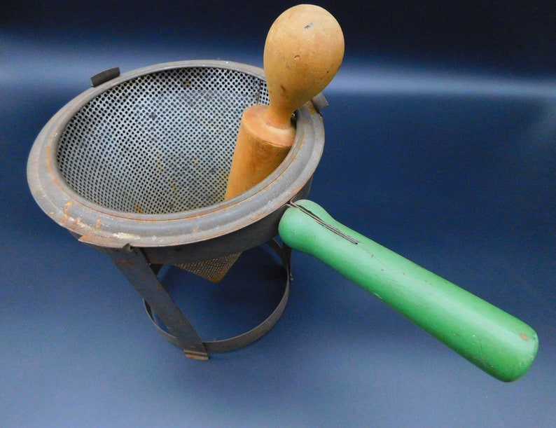 May include: A vintage metal food mill with a green wooden handle and a wooden pestle. The mill has a mesh strainer and a metal base.