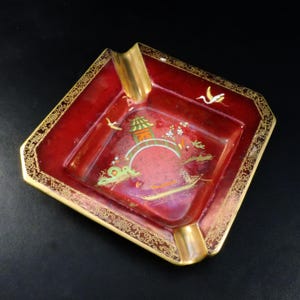 May include: A red and gold ashtray with an Asian-inspired design. The square ashtray features a gold rim and two gold rests. The center has a painted scene with a bridge, pagoda, and birds.