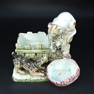 May include: A ceramic planter with a white cat figurine perched on a stone wall, a brown and white dog figurine peeking out from behind a green fence, and a pink and white bowl with a blue interior.