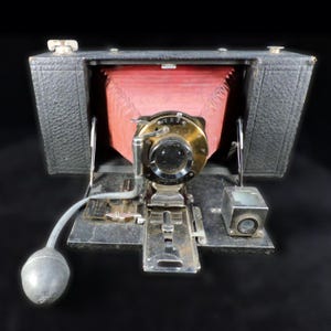 May include: Vintage black and silver folding camera with a red bellows. The camera has a brass lens and a silver viewfinder. A gray bulb is connected to the camera. The camera is open and ready to use.