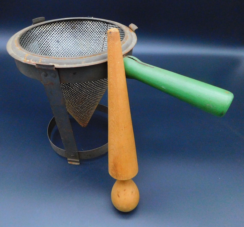May include: A vintage metal food mill with a green wooden handle and a wooden potato masher. The food mill has a conical shape with a mesh screen and a metal base.