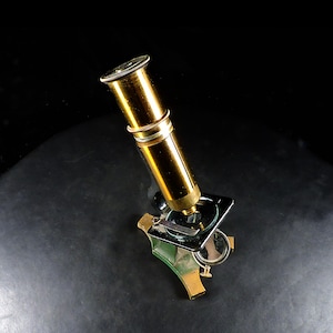 May include: A vintage brass microscope featuring a cylindrical body, a black square stage with a metal clip, and a circular mirror attached to the base. The instrument has a polished metallic finish and a sturdy tripod-style stand for stability.