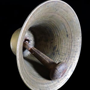 May include: Close-up of a bronze bell with a dark brown clapper. The bell has a wide, flared opening and a rounded top. The clapper is attached inside the bell and is designed to strike the inner surface to produce sound. The bell is set against a black background.