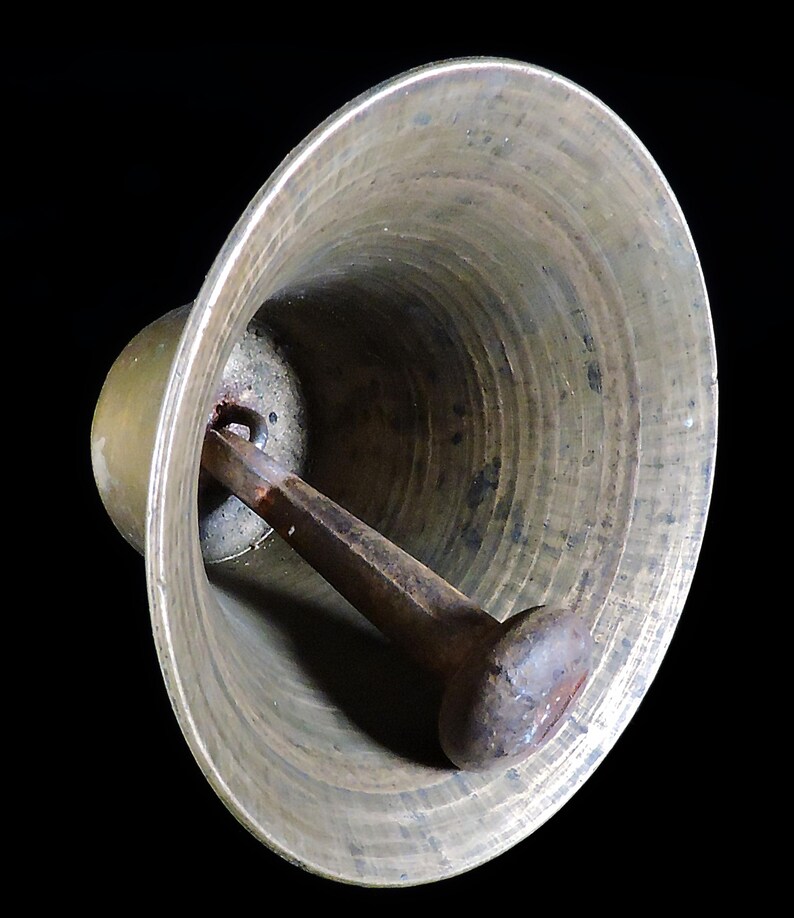 May include: Close-up of a bronze bell with a dark brown clapper. The bell has a wide, flared opening and a rounded top. The clapper is attached inside the bell and is designed to strike the inner surface to produce sound. The bell is set against a black background.