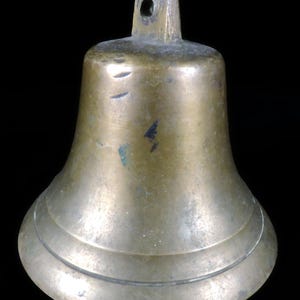 May include: A bronze bell with a flared base and a loop at the top for hanging. The bell has a slightly tarnished appearance, with some darker spots and scratches visible on the surface. The bell is a classic shape, suitable for various uses.