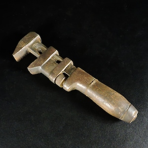 May include: A vintage adjustable wrench featuring a weathered metal head with a visible knurled adjustment nut and a worn, dark brown wooden handle. The tool displays signs of age and heavy use, with a textured, oxidized surface finish throughout.