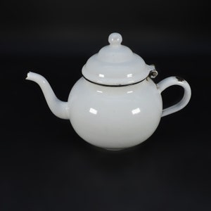 Peut inclure: Théière en émail blanc avec un couvercle et une poignée. La théière a un corps rond et un petit bec.